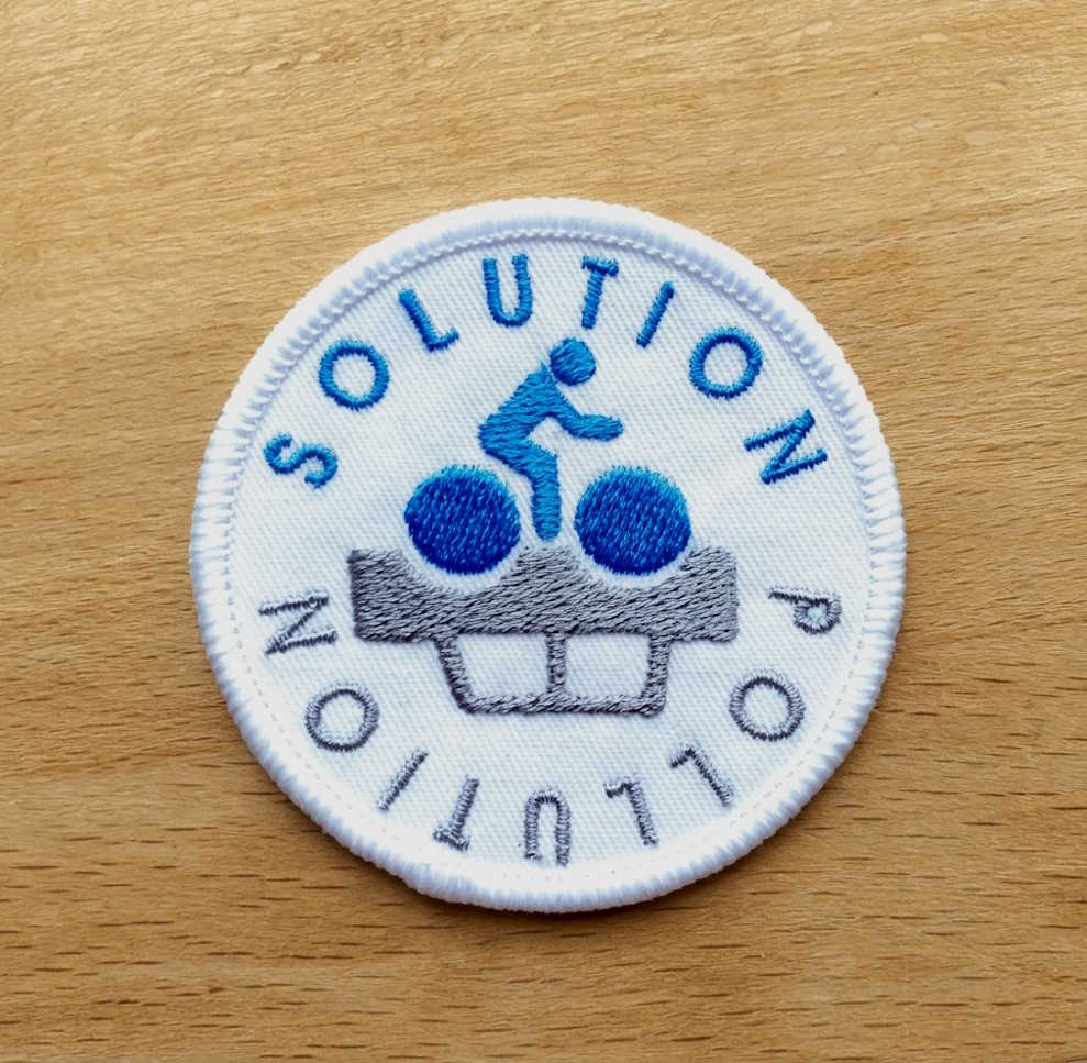 A circular embroidered patch on a light wood surface. The patch is white with blue and gray embroidery featuring the words 'SOLUTION POLLUTION' and a blue figure sitting on bicycle.