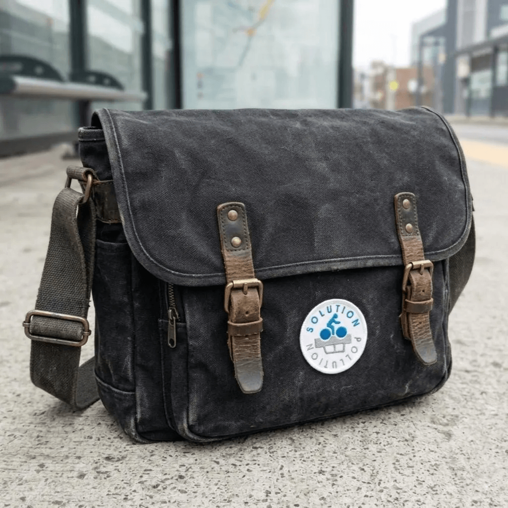 A dark gray washed canvas messenger bag with brown leather straps and a round white 'Solution Pollution' patch, shown on a concrete sidewalk with a blurred city street background.